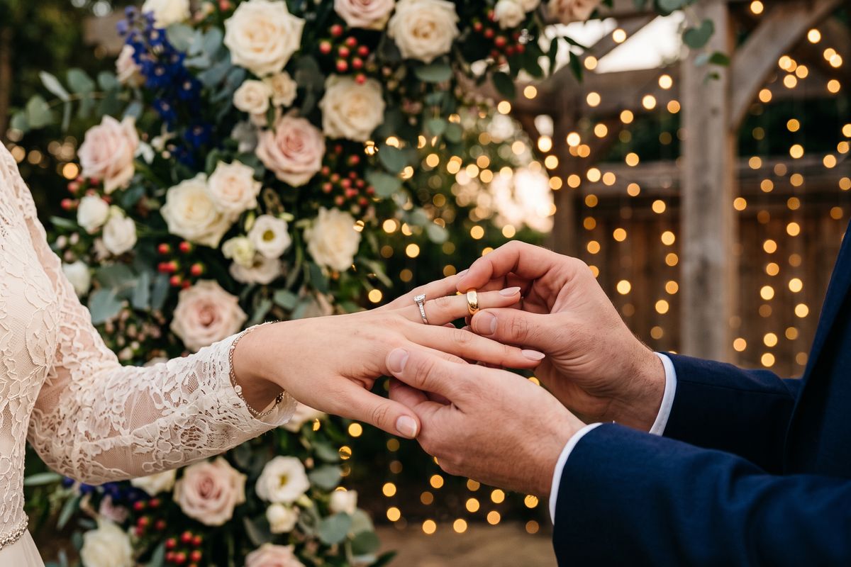 Groom placing a gold wedding ring on the bride's finger in front of a flower arch